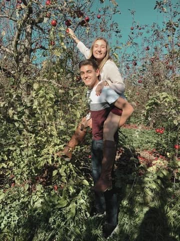 Allie and Alex apple picking in an orchard, Allie riding on Alex's back and reaching up toward the fruit.