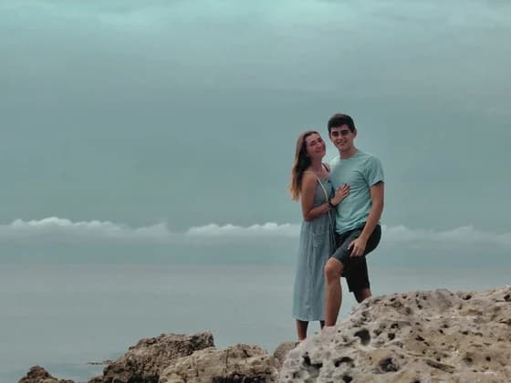 Allie and Alex standing together on pale rock at the edge of a flat grey sea, under heavy overcast.
