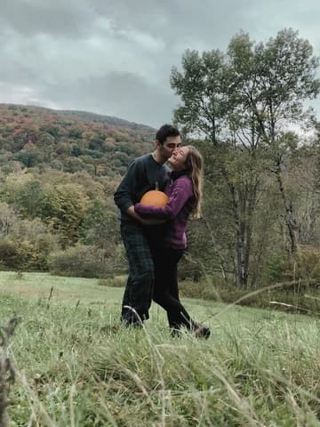 Allie and Alex standing in a mown field holding a pumpkin between them, forested hills turning colour behind.