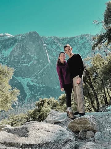 Allie and Alex standing on granite boulders, a rounded granite dome and conifer slopes behind them.