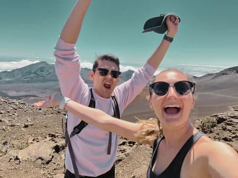 Allie and Alex taking a selfie with their arms raised on a bare volcanic crater rim above a layer of cloud.