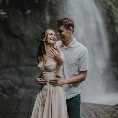 Allie and Alex in front of a waterfall, Allie in a pale gown with her hand raised to show her engagement ring.