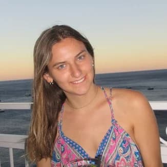 Bridesmaid: a woman with long light-brown hair in a pink and blue patterned dress on a balcony, the sea and a sunset sky behind her.