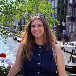 Bridesmaid: a woman with long light-brown hair in a denim waistcoat beside a canal, moored boats and brick townhouses behind her.