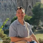 Groomsman: a man in a pale grey polo sitting on a stone wall with his arms folded, a large ornate basilica behind him.