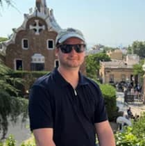 Groomsman: a man in a navy polo, sunglasses and a cap sitting on a curved tiled wall, mosaic-roofed pavilions behind him.